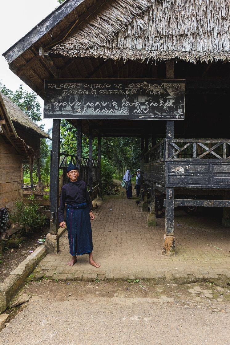 A Kajang elder in traditional black dress beneath the customary welcome sign at the entrance to Tana Toa